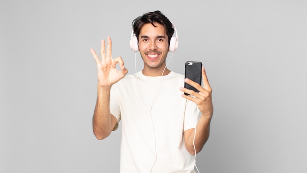 Jovem hispânico se sentindo feliz, mostrando aprovação com um gesto de ok com fones de ouvido e smartphone