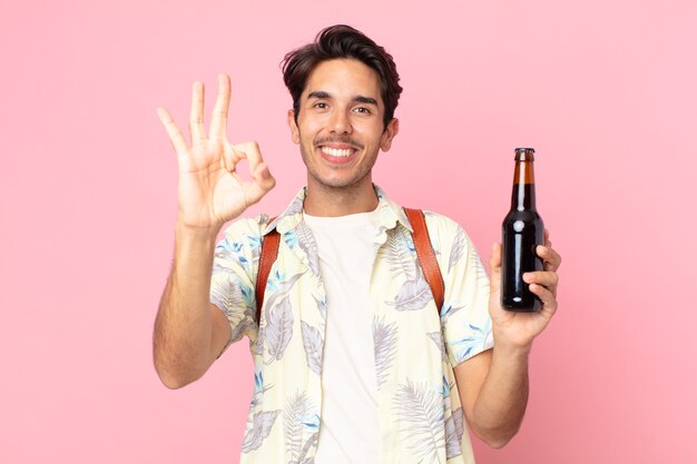 Jovem hispânico se sentindo feliz, mostrando aprovação com um gesto de ok e segurando uma garrafa de cerveja