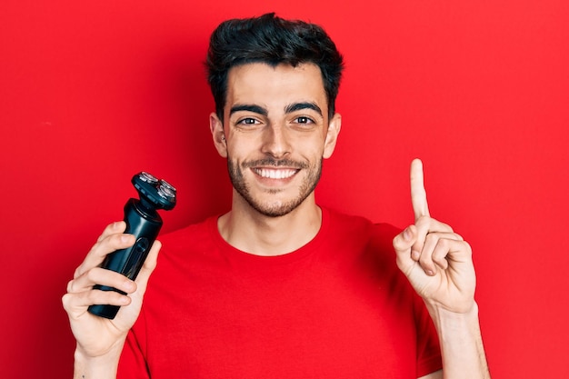 Jovem hispânico segurando a máquina de barbear elétrica sorrindo com uma ideia ou pergunta apontando o dedo com cara feliz número um