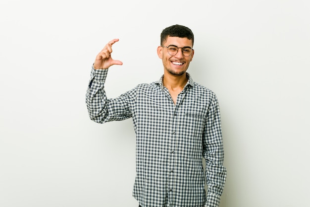 Jovem hispânico segurando algo pequeno com os indicadores, sorrindo e confiante.