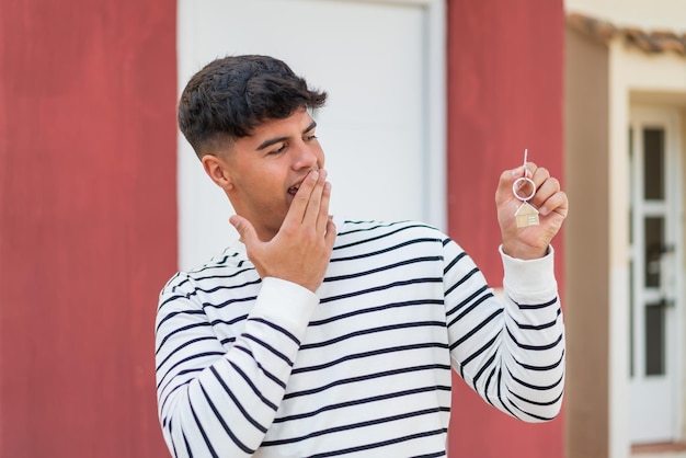 Jovem hispânico segurando chaves de casa ao ar livre com expressão facial de surpresa e choque