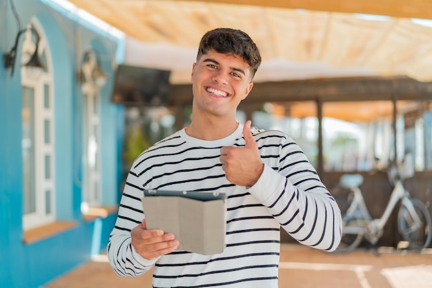 Foto jovem hispânico segurando um tablet ao ar livre com os polegares para cima porque algo bom aconteceu