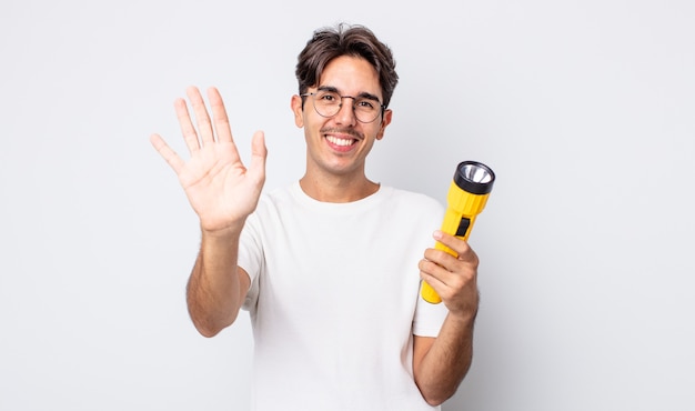 Jovem hispânico sorrindo alegremente, acenando com a mão, dando as boas-vindas e cumprimentando você. conceito de lanterna