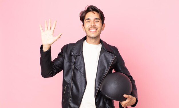 Jovem hispânico sorrindo alegremente, acenando com a mão, dando as boas-vindas e cumprimentando você. conceito de motociclista