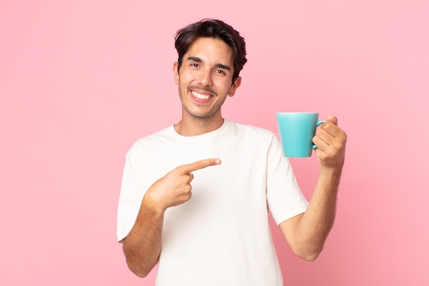 Jovem hispânico sorrindo alegremente, sentindo-se feliz, apontando para o lado e segurando uma caneca de café