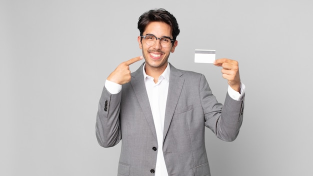 Jovem hispânico sorrindo com confiança, apontando para o próprio sorriso largo e segurando um cartão de crédito