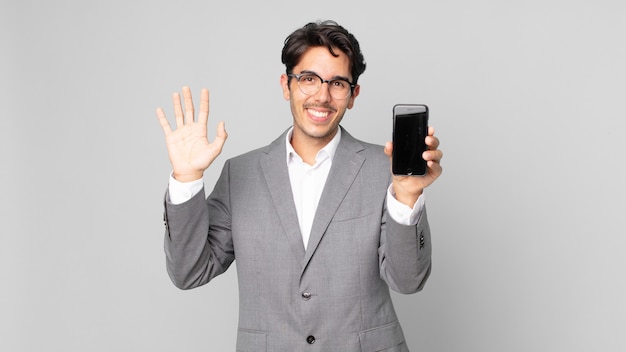 Jovem hispânico sorrindo feliz, acenando com a mão, dando as boas-vindas, cumprimentando você e segurando um smartphone