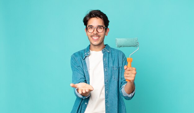 Jovem hispânico sorrindo feliz com simpatia e oferecendo e mostrando um conceito e segurando um rolo de pintura