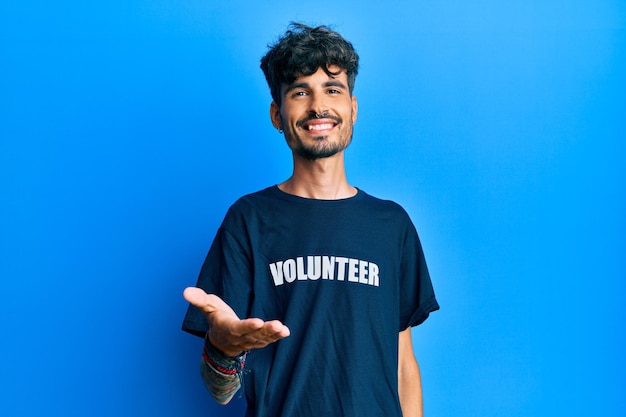 Jovem hispânico vestindo camiseta de voluntário sorrindo alegre oferecendo mão de palma dando assistência e aceitação.