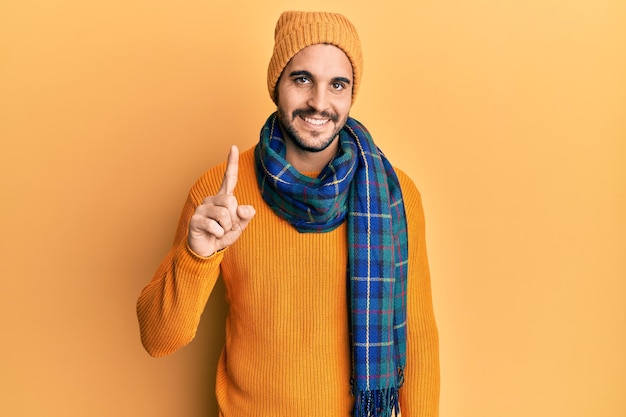 Jovem hispânico vestindo suéter de lã e cachecol de inverno sorrindo com uma ideia ou pergunta apontando o dedo para cima com cara feliz número um