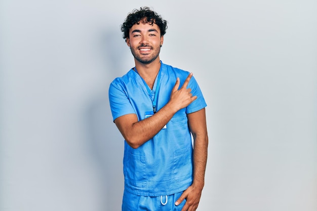 Jovem hispânico vestindo uniforme de enfermeiro azul alegre com um sorriso no rosto apontando com a mão e o dedo para o lado com expressão feliz e natural no rosto