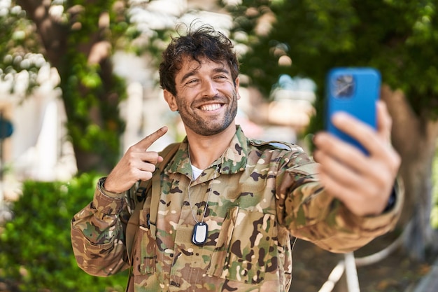 Jovem hispânico vestindo uniforme do exército de camuflagem fazendo videochamada sorrindo feliz apontando com a mão e o dedo
