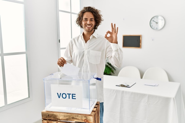 Foto jovem hispânico votando colocando envelope na urna sorrindo positivo fazendo sinal de ok com mão e dedos expressão de sucesso