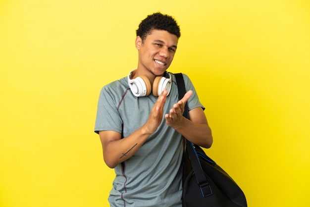 Jovem homem afro-americano com uma bolsa esportiva isolada em um fundo amarelo, aplaudindo após uma apresentação em uma conferência