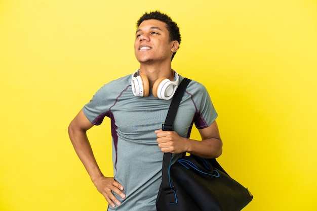 Jovem homem afro-americano com uma bolsa esportiva isolada em um fundo amarelo, posando com os braços no quadril e sorrindo