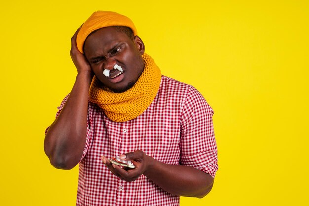 Jovem homem afro-americano vestindo camisa quadriculada vermelha com cachecol de malha e chapéu se sentindo mal com febre calor e calafrios no estúdio amarelo background.flu e conceito de vírus.