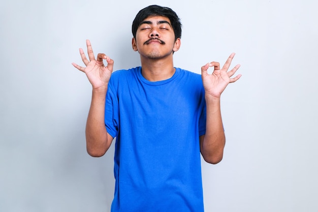 Jovem homem asiático vestindo camiseta casual em pé sobre fundo branco isolado relaxar e sorrindo com os olhos fechados, fazendo o gesto de meditação com os dedos. conceito de ioga.