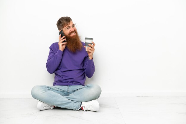 Jovem homem avermelhado, caucasiano, sentado no chão, isolado no fundo branco, segurando um café para levar e um celular