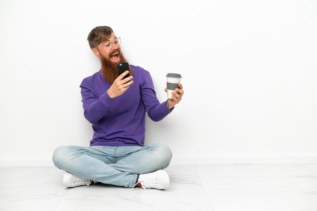 Jovem homem avermelhado, caucasiano, sentado no chão, isolado no fundo branco, segurando um café para levar e um celular