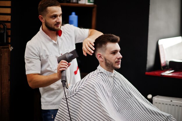 Jovem homem barbudo cortando cabelo de cabeleireiro enquanto está sentado na cadeira de barbearia. Alma de barbeiro.