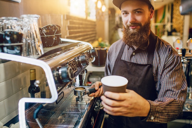 Jovem homem barbudo fica na máquina de café e mantém a xícara de café com leite na mão