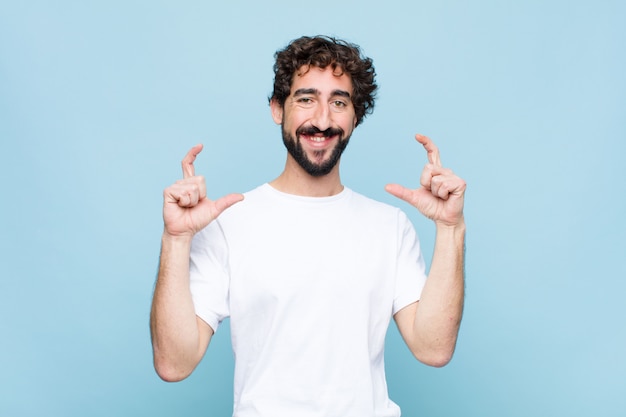 Jovem homem barbudo louco emoldurando ou descrevendo o próprio sorriso com as duas mãos, olhando positivo e feliz, conceito de bem-estar contra a parede plana