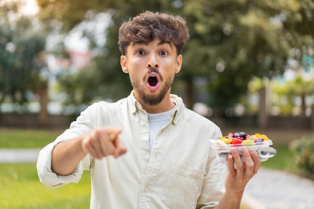 Foto jovem homem bonito árabe segurando uma tigela de frutas ao ar livre surpreso e apontando para a frente