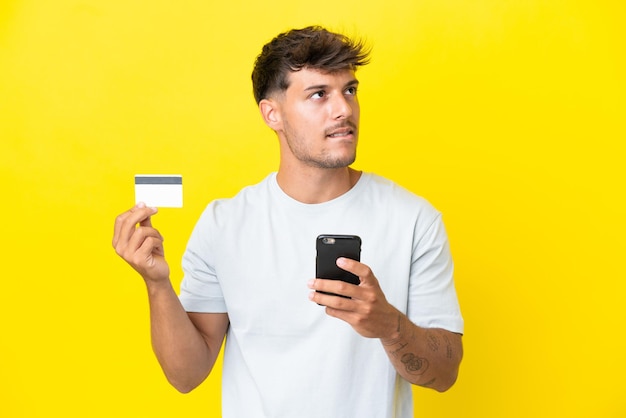 Jovem homem bonito, caucasiano, isolado em um fundo amarelo, comprando com o celular com um cartão de crédito enquanto pensa