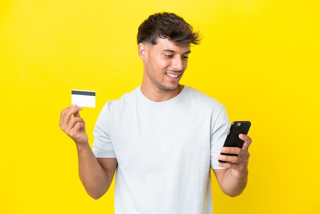 Jovem homem bonito, caucasiano, isolado em um fundo amarelo, comprando com o celular com um cartão de crédito