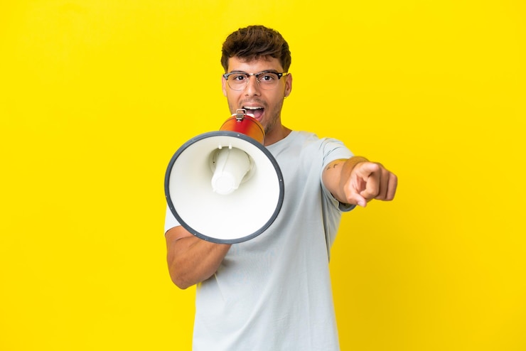 Foto jovem homem bonito, caucasiano, isolado em um fundo amarelo, gritando em um megafone para anunciar algo enquanto aponta para a frente