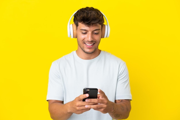 Jovem homem bonito, caucasiano, isolado em um fundo amarelo, ouvindo música e olhando para o celular