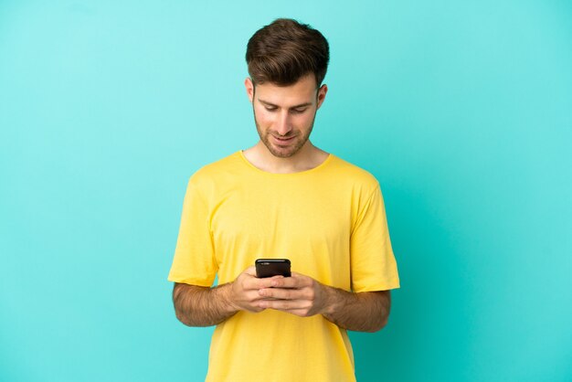 Jovem homem bonito, caucasiano, isolado em um fundo azul, enviando uma mensagem com o celular