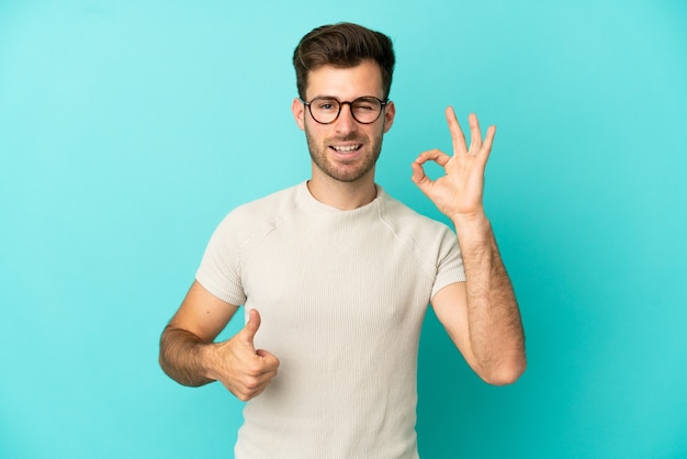 Jovem homem bonito, caucasiano, isolado em um fundo azul, mostrando sinal de ok e gesto de polegar para cima