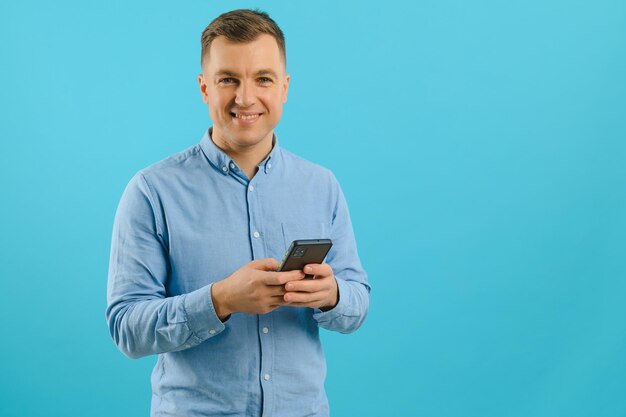 Jovem homem bonito, caucasiano, isolado em um fundo azul, olhando para a câmera e sorrindo enquanto usa o celular