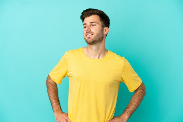 Jovem homem bonito, caucasiano, isolado em um fundo azul, posando com os braços na cintura e sorrindo