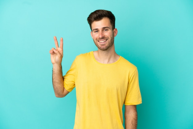 Jovem homem bonito, caucasiano, isolado em um fundo azul, sorrindo e mostrando sinal de vitória