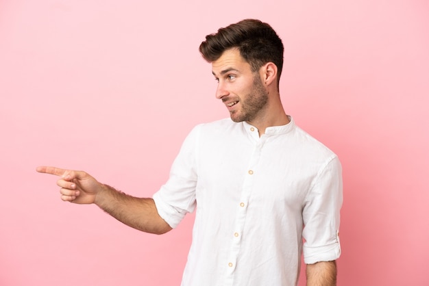 Jovem homem bonito, caucasiano, isolado em um fundo rosa, apontando o dedo para o lado e apresentando um produto