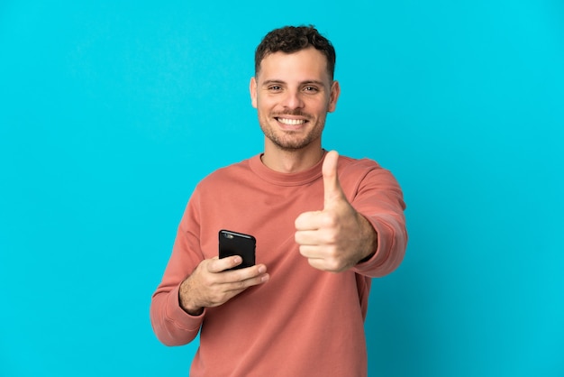 Jovem homem bonito, caucasiano, isolado na parede azul, usando um telefone celular enquanto faz o polegar para cima