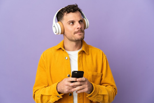 Jovem homem bonito, caucasiano, isolado na parede roxa, ouvindo música com um celular e pensando