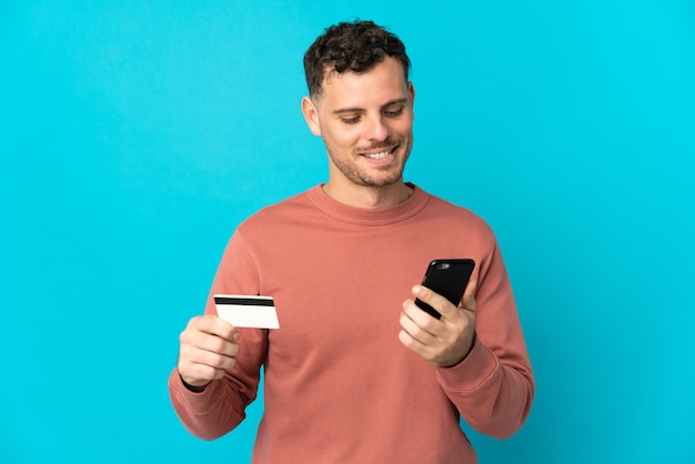 Jovem homem bonito, caucasiano, isolado no azul, comprando com o celular com um cartão de crédito