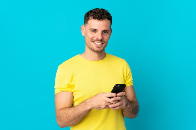 Jovem homem bonito, caucasiano, isolado no azul, enviando uma mensagem com o celular