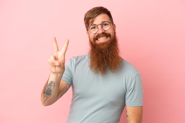 Jovem homem branco avermelhado isolado em um fundo rosa sorrindo e mostrando sinal de vitória