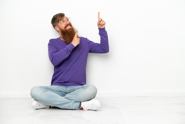 Jovem homem branco avermelhado sentado no chão isolado no fundo branco apontando com o dedo indicador uma ótima ideia