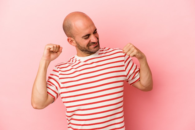 Jovem homem careca caucasiano isolado em um fundo rosa, comemorando um dia especial, pula e levanta os braços com energia.