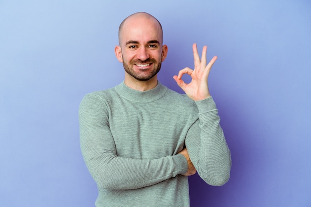 Jovem homem careca caucasiano isolado em um fundo roxo pisca um olho e segura um gesto de ok com a mão.