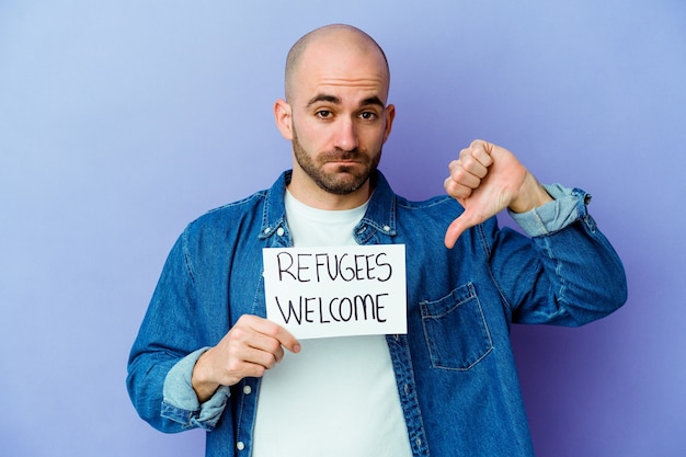 Jovem homem careca, caucasiano, segurando um cartaz de boas-vindas aos refugiados, isolado em um fundo azul, mostrando um gesto de antipatia, polegares para baixo. Conceito de desacordo.