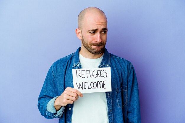 Jovem homem careca, caucasiano, segurando um cartaz de boas-vindas aos refugiados, isolado na parede azul, confuso, sente-se em dúvida e inseguro.