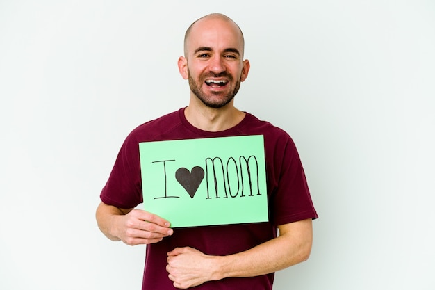 Jovem homem careca, caucasiano, segurando um cartaz de eu amo a mãe, isolado na parede amarela, rindo e se divertindo