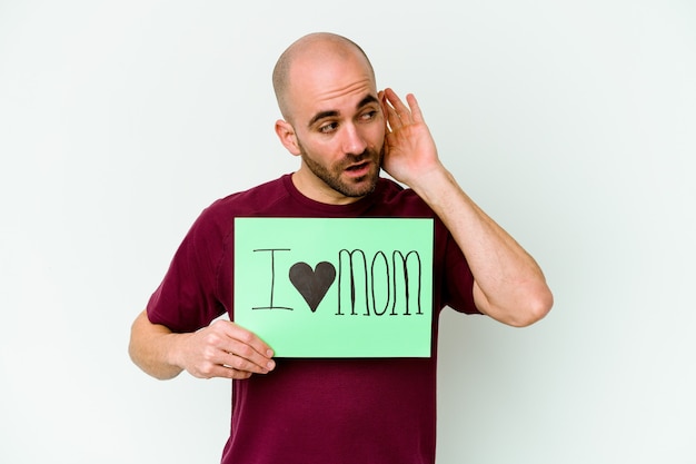 Jovem homem careca caucasiano segurando um cartaz de eu amo mãe isolado na parede amarela, tentando ouvir uma fofoca.