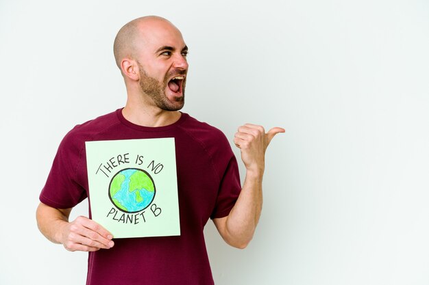 Jovem homem careca caucasiano segurando um cartaz de não há planeta B isolado na parede roxa aponta com o dedo polegar afastado, rindo e despreocupado.